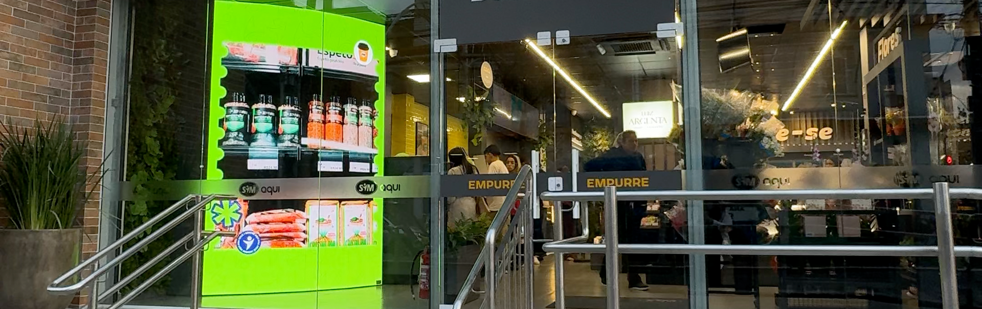 Painel de LED vertical instalado na entrada de uma loja de conveniência, exibindo produtos e promoções digitais em alta definição. O painel está integrado à fachada de vidro do estabelecimento, destacando-se pela luminosidade e pelas cores vibrantes, atraindo a atenção dos clientes que circulam pelo local.